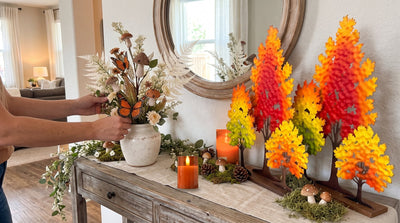 Two Triple Fall Trees, two orange glass flameless candles and a vase of mushroom spray and white fern on a console table