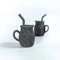 Two black ceramic mugs with straw holders on a white background