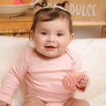 Baby in a pink outfit holding a toy, sitting on a white surface with a blurred background.