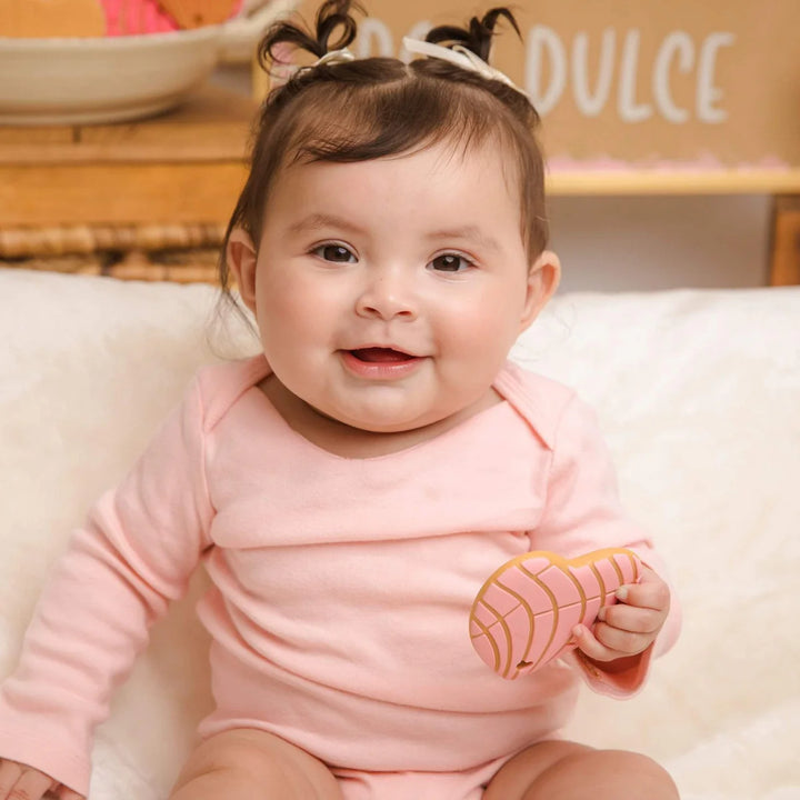 Baby in a pink outfit holding a toy, sitting on a white surface with a blurred background.