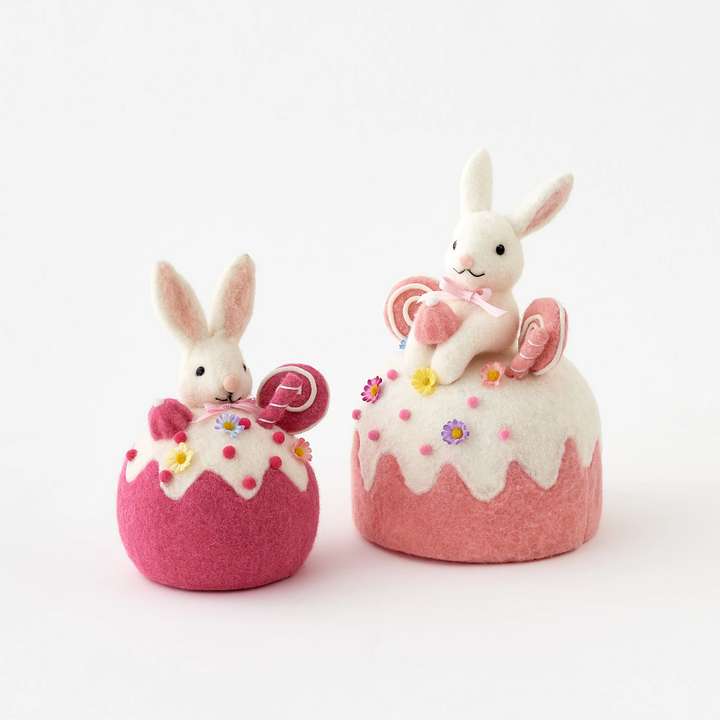 Two felted rabbit toys on pink cake-shaped bases with white frosting on a white background