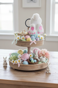 Two-tiered wooden tray with Easter decorations including eggs, flowers, and bunnies on a table.
