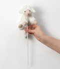 Hand holding a fluffy white toy sheep on a stick against a plain background
