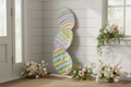 Decorative wall art with colorful Easter egg design in a room with flowers and a window.